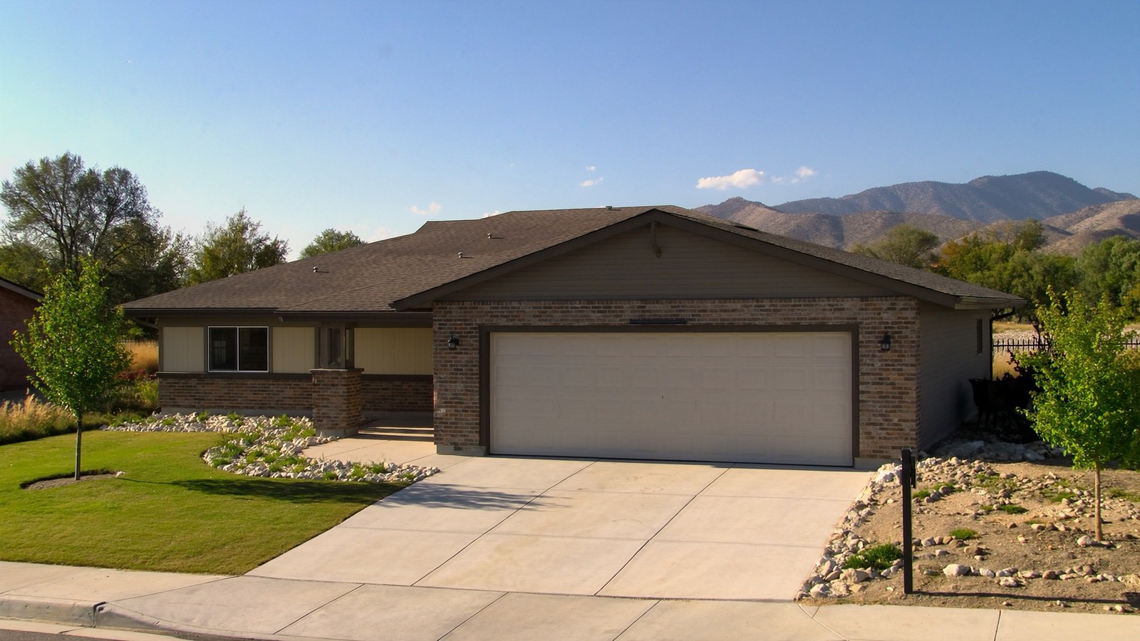 A one-story brick and siding ranch-style home in a Salt Lake Valley suburb, with a mixed lawn and xeriscape front yard, an attached two-car garage, and the Wasatch Range foothills in the background